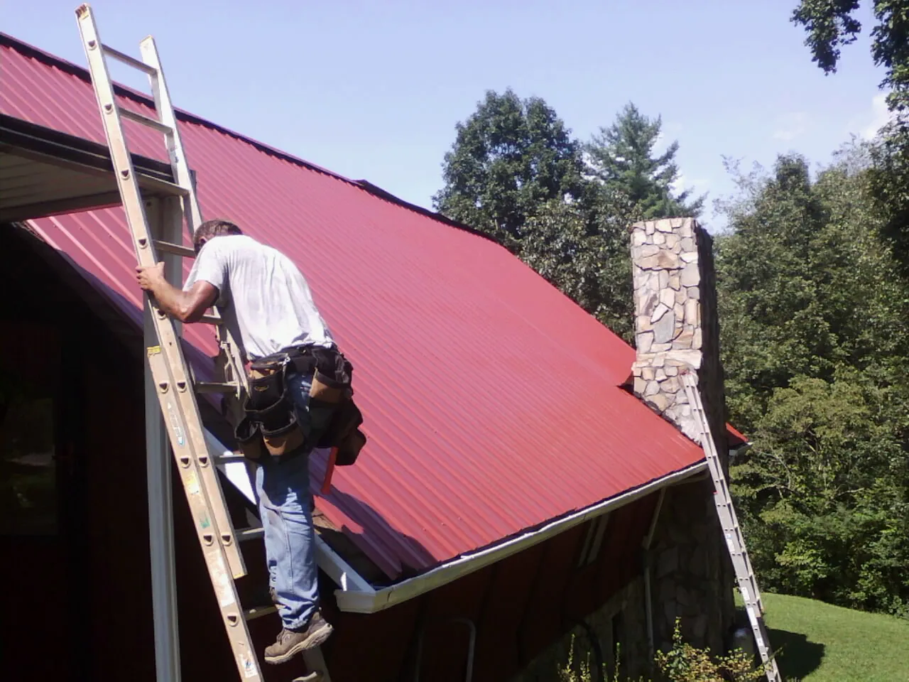 Roofer on a ladder beside a red metal roof and stone chimney — Soffit & Fascia Repair by Roofing Brothers in Moody